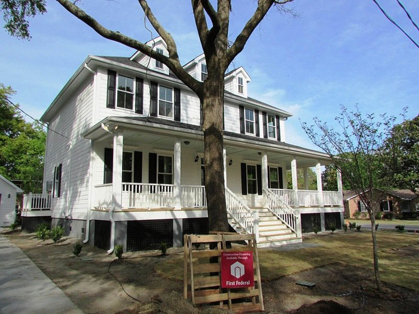 White two-story house with wide front porch, large tree with leafy canopy in landscaped yard, red real estate sign on wooden rack near walkway, multiple windows and light siding