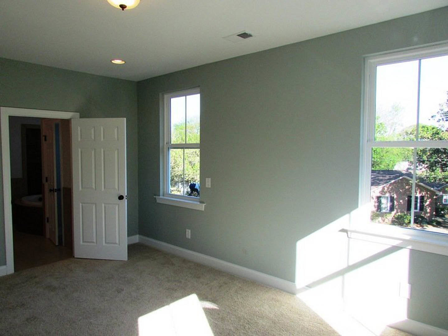 Open white door with black handle leading into a room, window with tree view, light fixture on ceiling, neutral walls and flooring