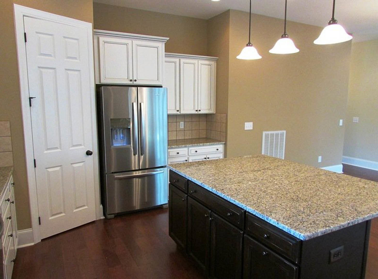 Granite island with blue speckled countertop, stainless steel refrigerator, white cabinetry, ceiling pendant lights, white door with black handle, and vent above sink