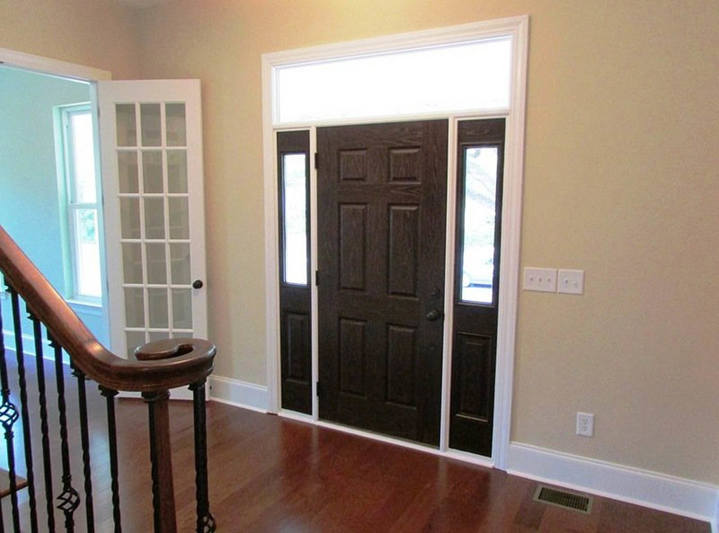 Dark brown wood door with brushed metal handle, white wall with electrical outlet and air vent, laminate flooring, staircase partially visible in background