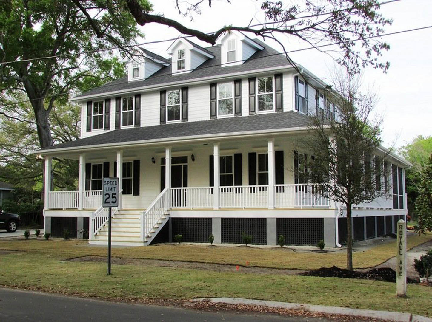 White two-story house with wide covered porch, white siding, black porch columns, white framed windows, and front staircase; speed limit sign and tree in foreground.