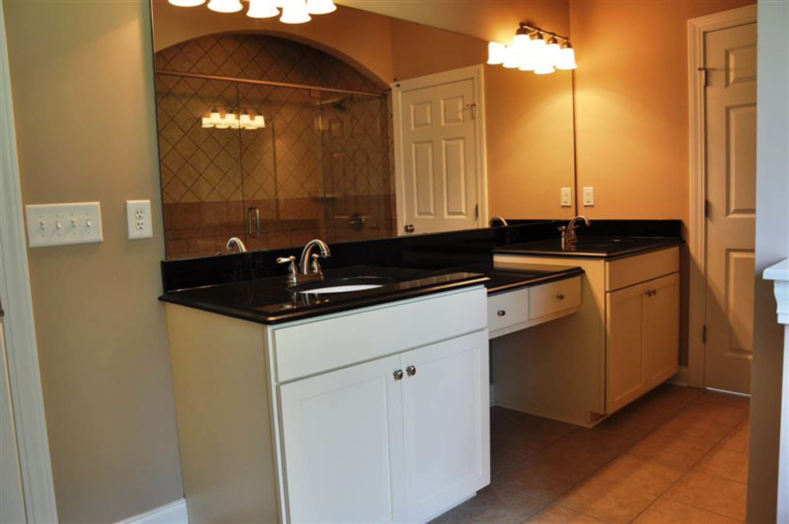 Bathroom with a large frameless mirror above a white vanity cabinet, black stone countertop, modern sink and faucet, glass shower wall, white paneled door, and tiled walls