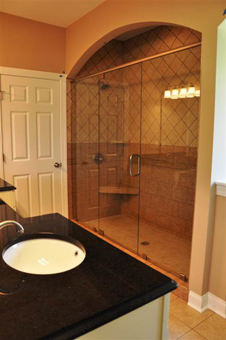 Modern bathroom featuring a frameless glass shower enclosure, white countertop sink with chrome faucet, light tile flooring, and a frosted window providing natural light.