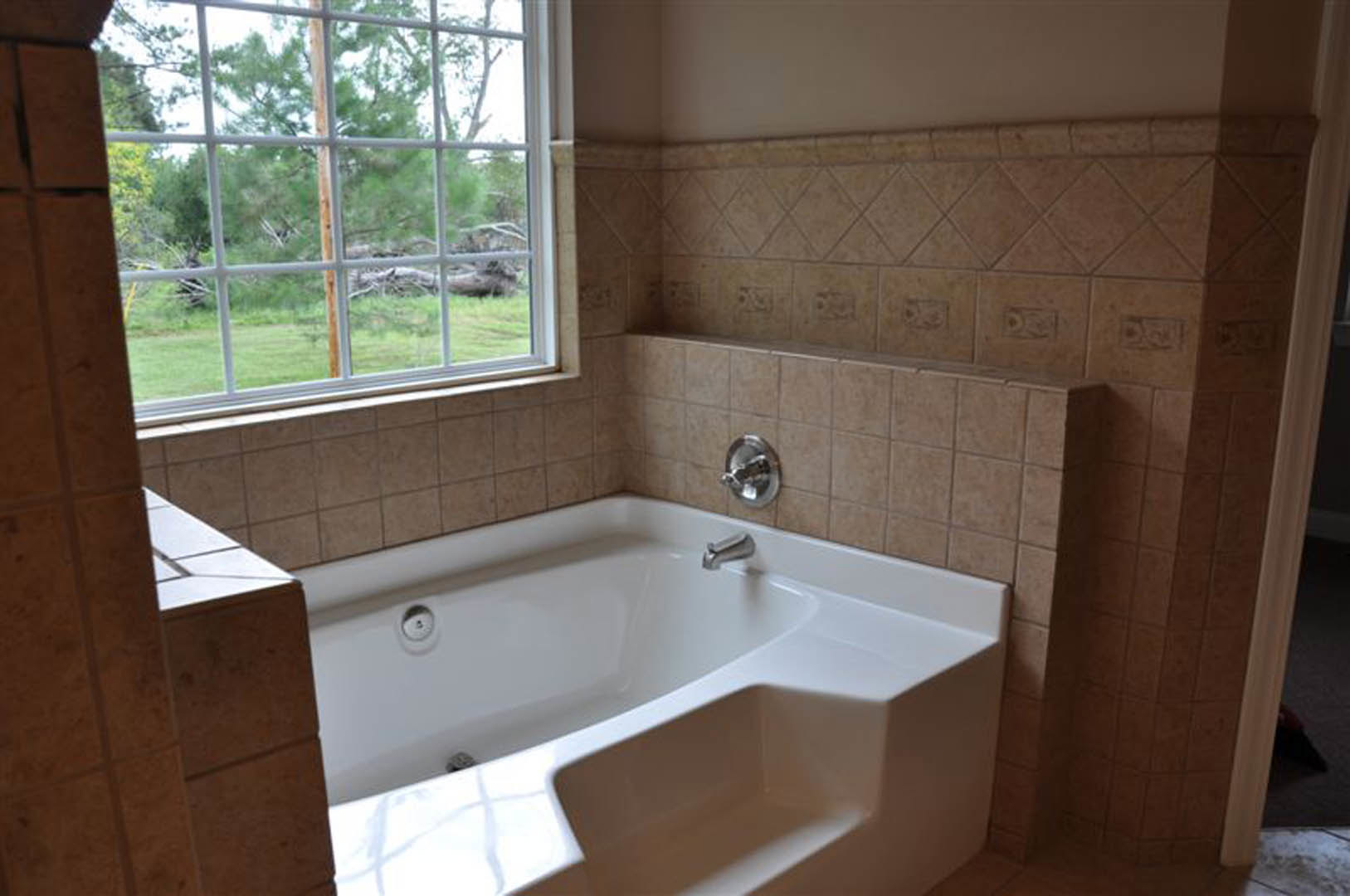 Freestanding white bathtub with chrome faucet on tiled floor, large window with natural light, neutral wall tiles, modern bathroom accessories