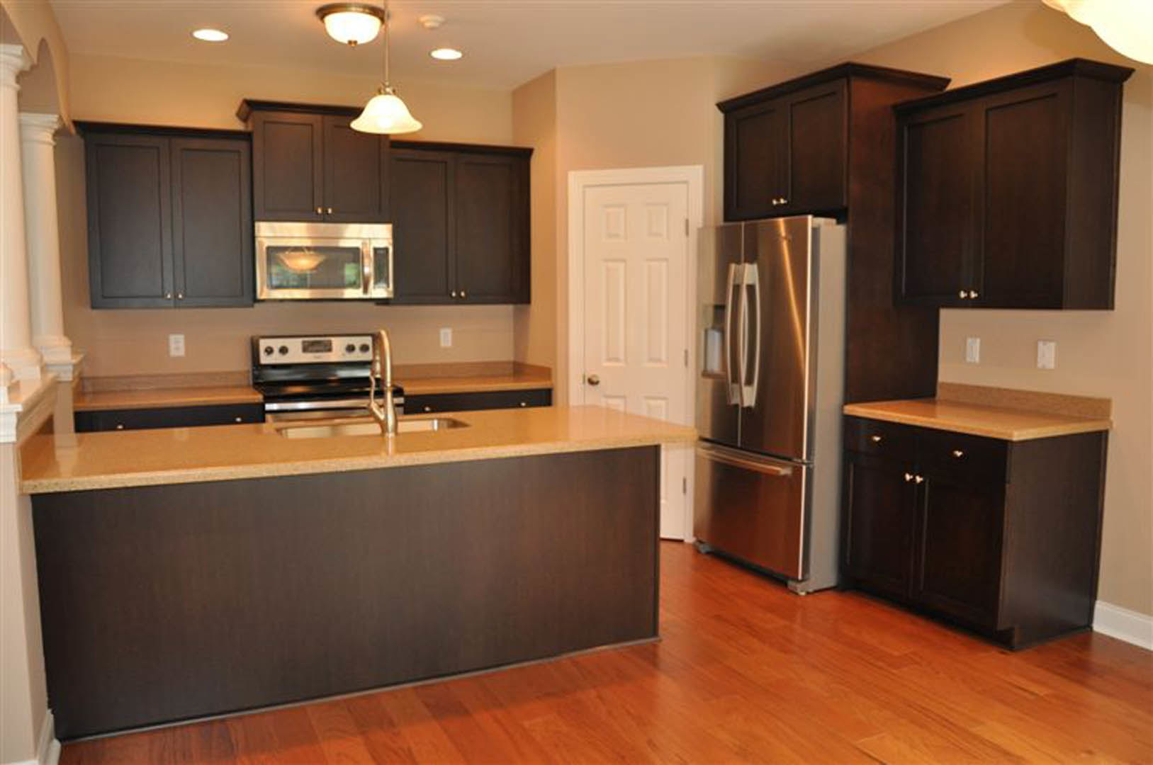 Dark wood kitchen cabinets, stainless steel refrigerator, tan countertop with sink, oven, and blurred light fixture above