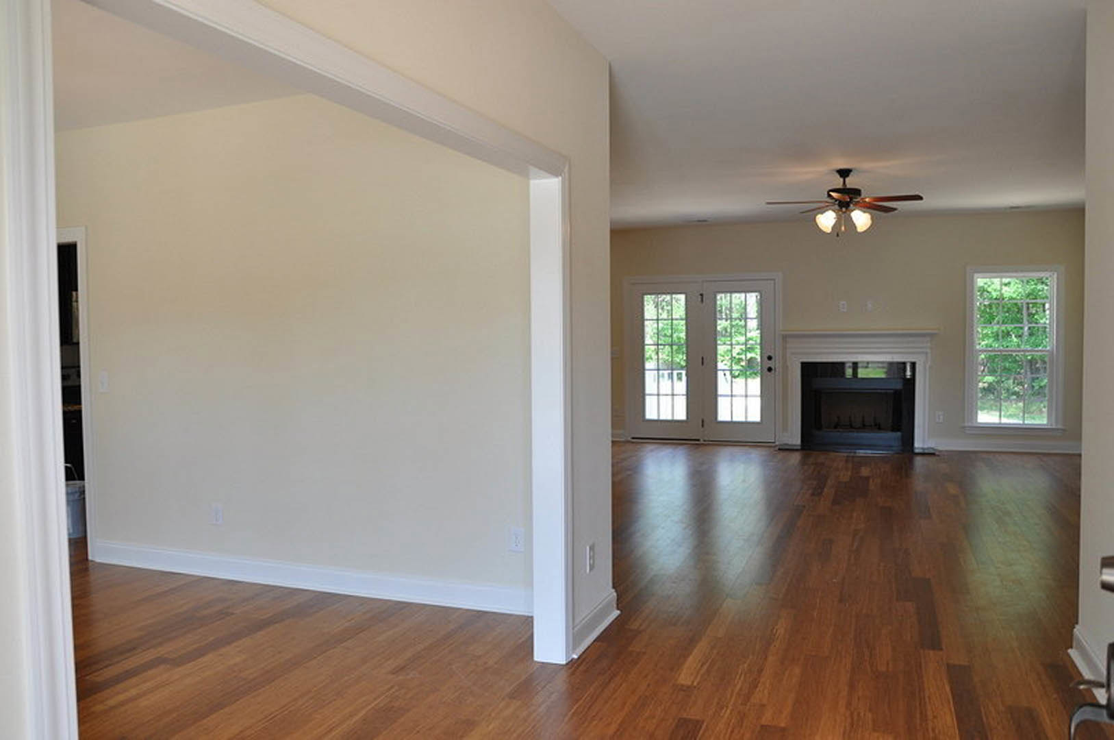 Living room with hardwood flooring, ceiling fan with lights, glass-paneled double doors, white column, white-framed fireplace, and window overlooking trees.