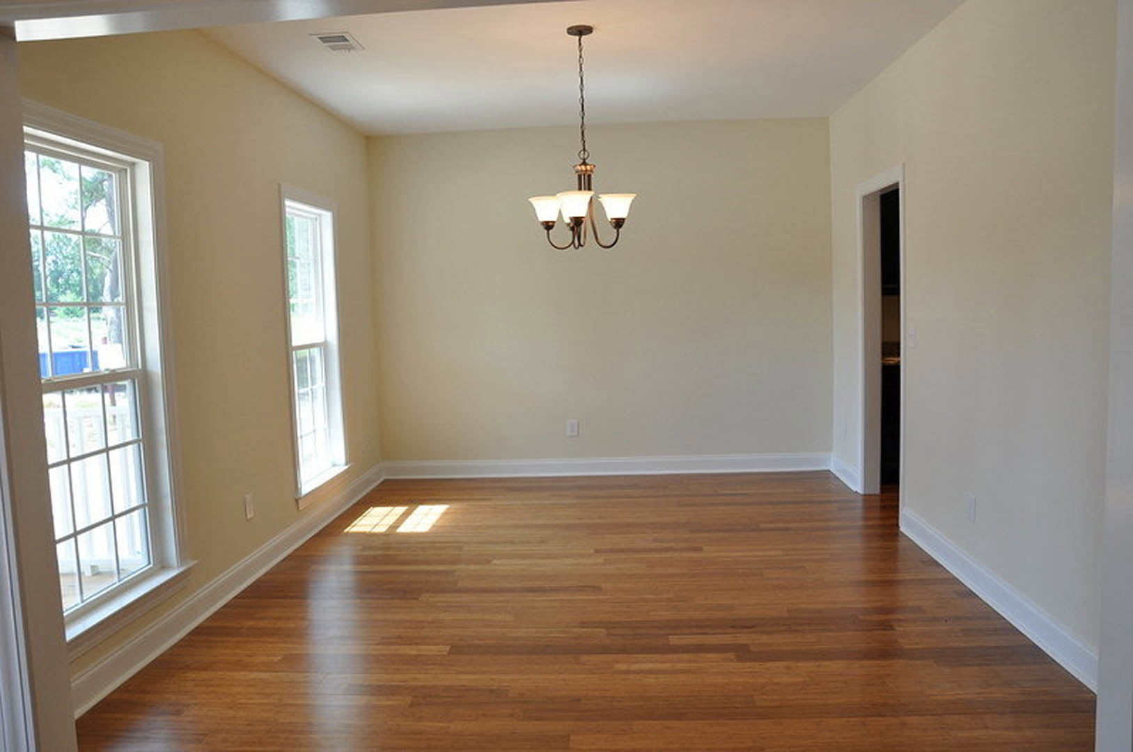 Chandelier hanging in a room with hardwood floors, sunlight streaming through a large white-framed window, plaster walls, and wood finishes.
