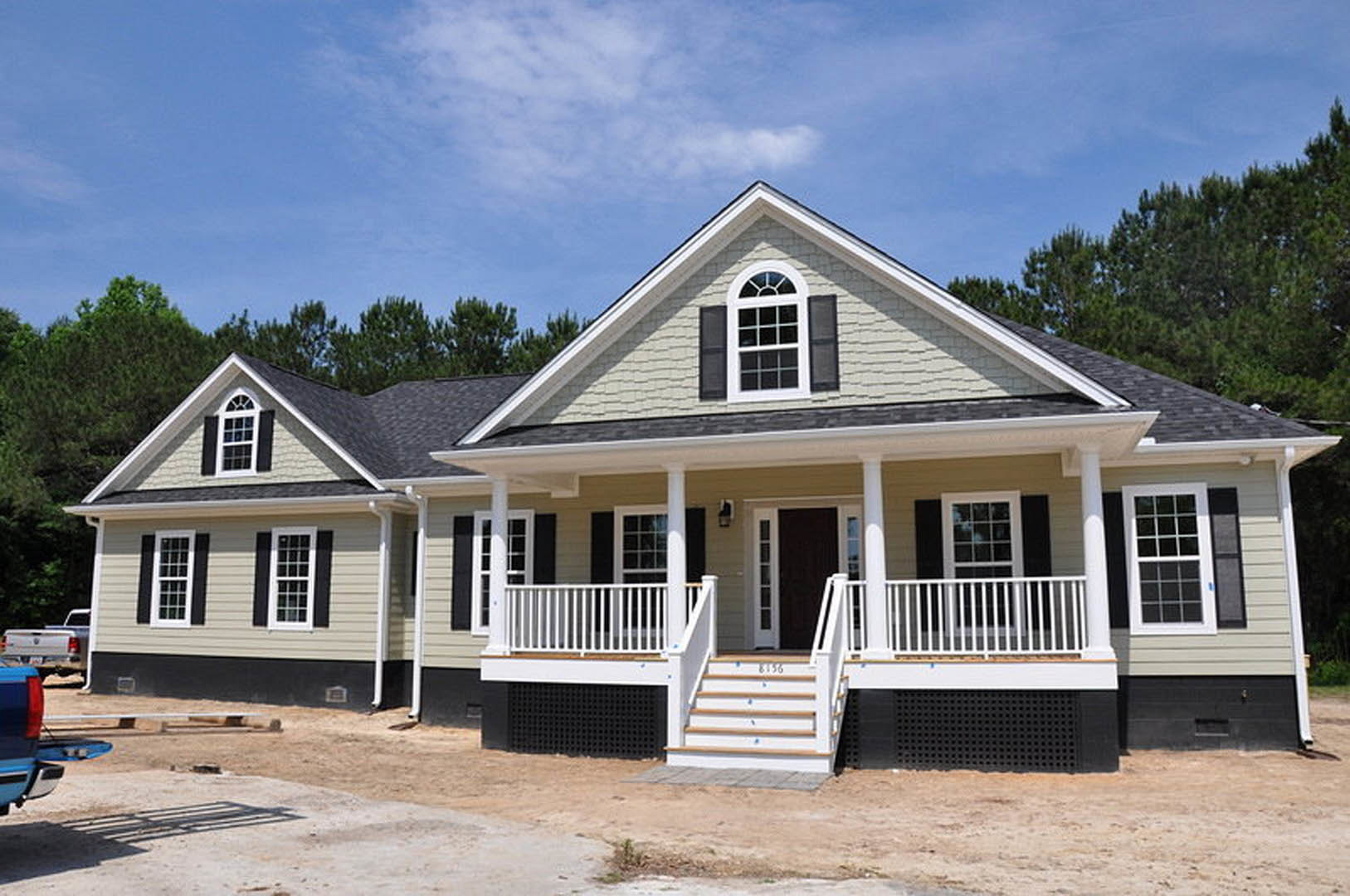 Two-story house under construction with exposed framing, white porch columns, multi-pane windows, and a white staircase, set against a clear blue sky.