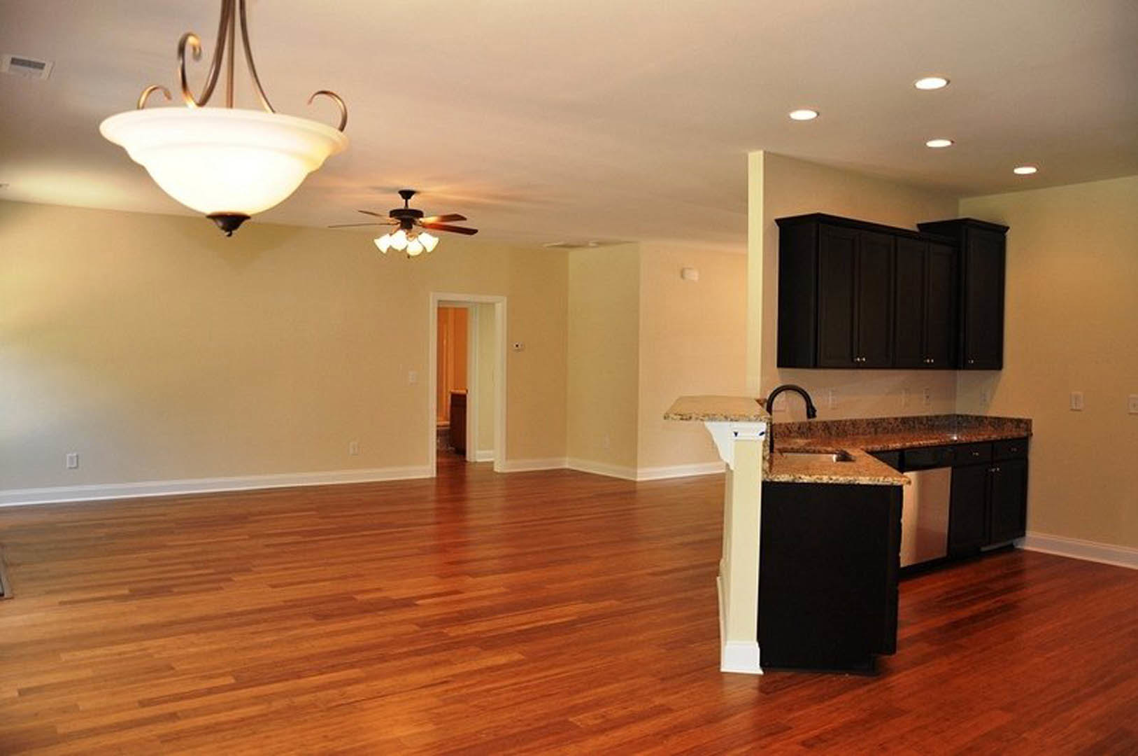 Open-concept kitchen and living room featuring hardwood flooring, black cabinetry, light fixtures, and wood-edged accents with natural sunlight illuminating the space.