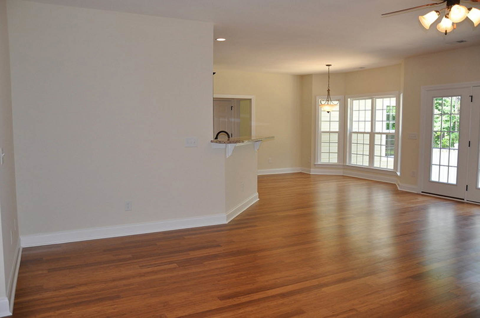 Empty living room with hardwood floors, white plaster walls, large window overlooking trees, modern light fixture, and wooden table with a long stick placed on top