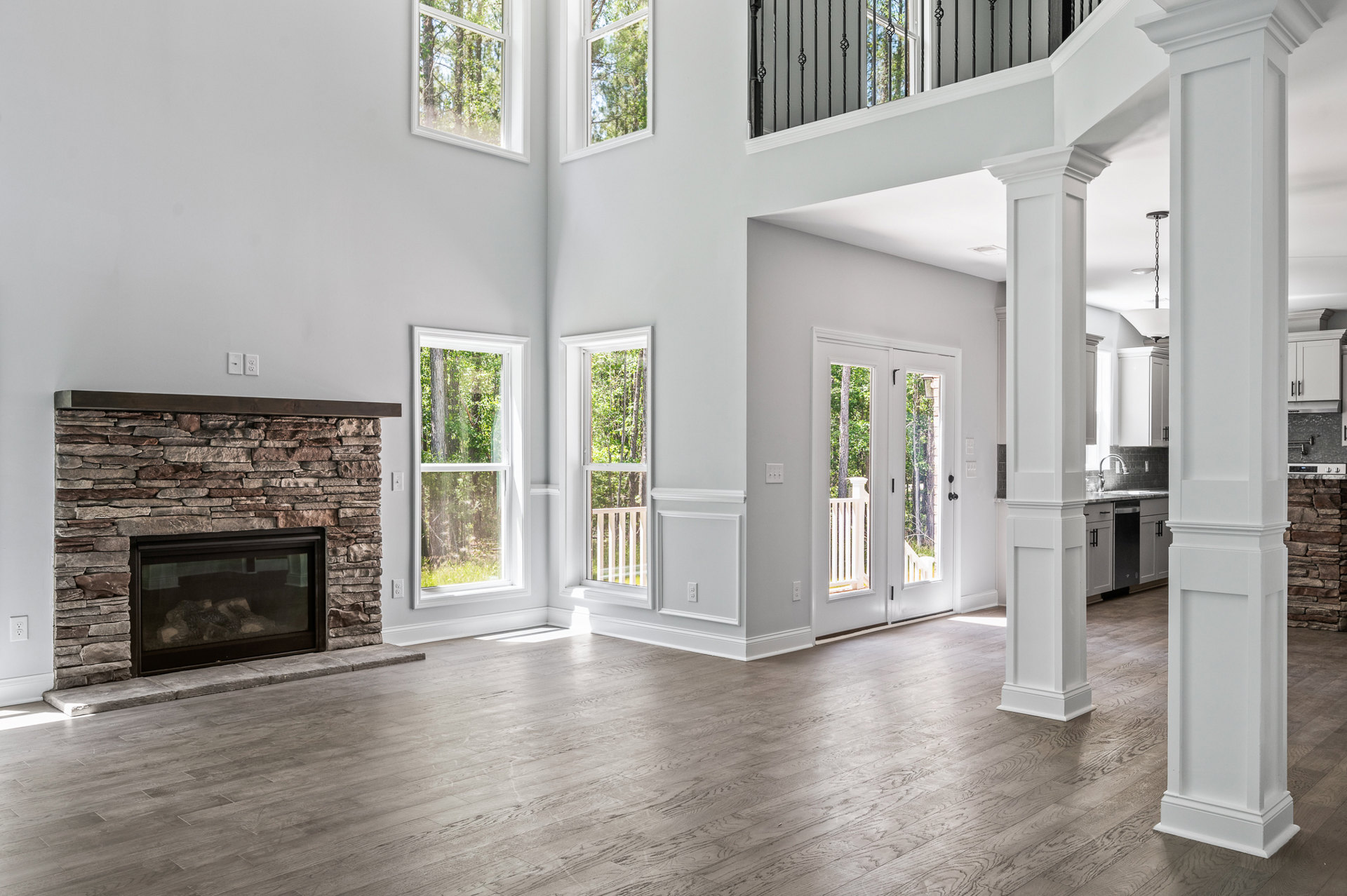 Spacious living room featuring a glass-door fireplace set into a large white wall, wood flooring, tall windows with tree views, and a white pillar visible near the kitchen area