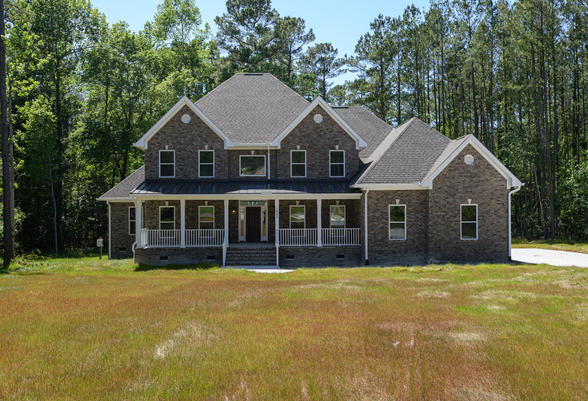 Two-story house with white porch railing, black front door, large windows, and expansive green lawn bordered by trees under a blue sky