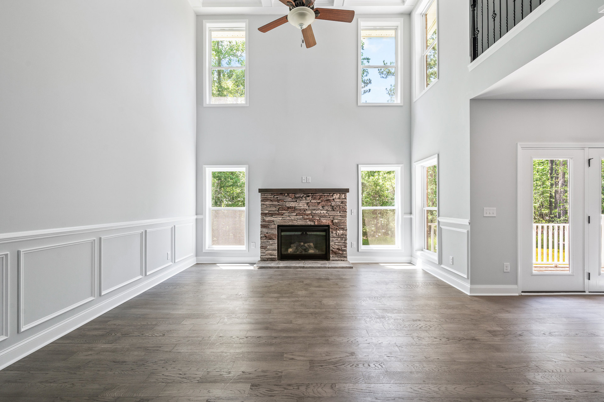 Living room with stone fireplace, firewood stacked inside, wood flooring, ceiling fan with light fixture, large window overlooking trees