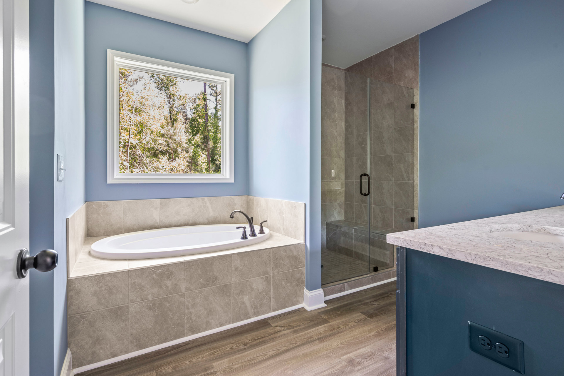Bathroom with freestanding bathtub and glass-enclosed shower, white tile walls, large window framing trees, quartz countertop with undermount sink and chrome faucet, electrical