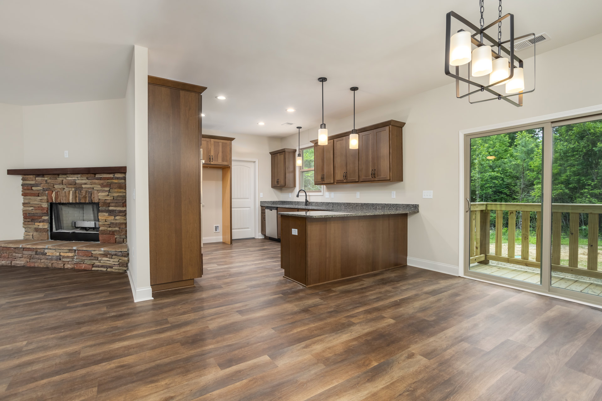 Open kitchen and living room featuring wood flooring, four-lamp chandelier, brick fireplace with black mesh cover, white brick accent wall, brown wooden door, and white cabinetry.