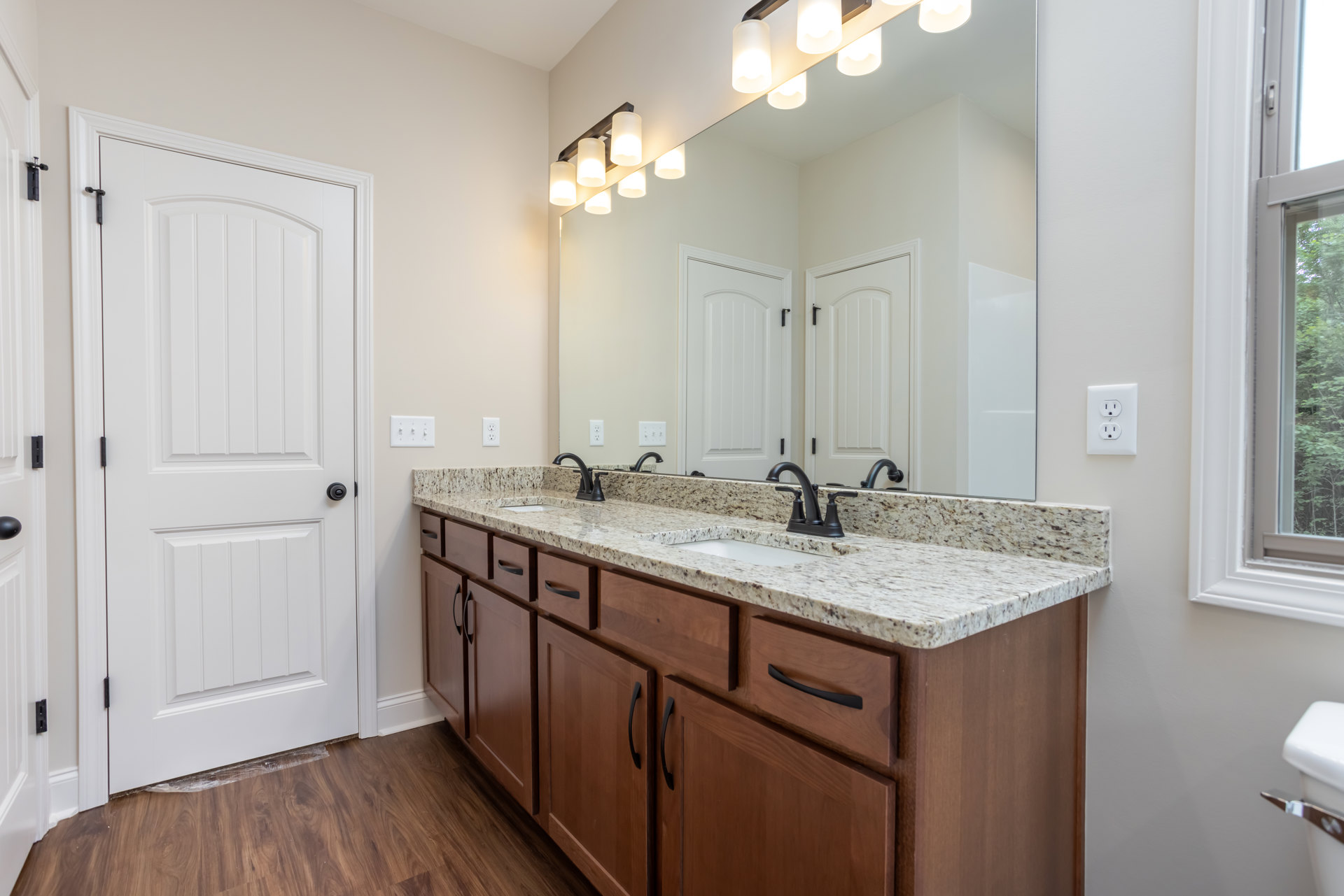 Double vanity bathroom with wooden cabinets, large wall mirror, white door with black handle, tile flooring, chrome faucet, and visible electrical outlet.