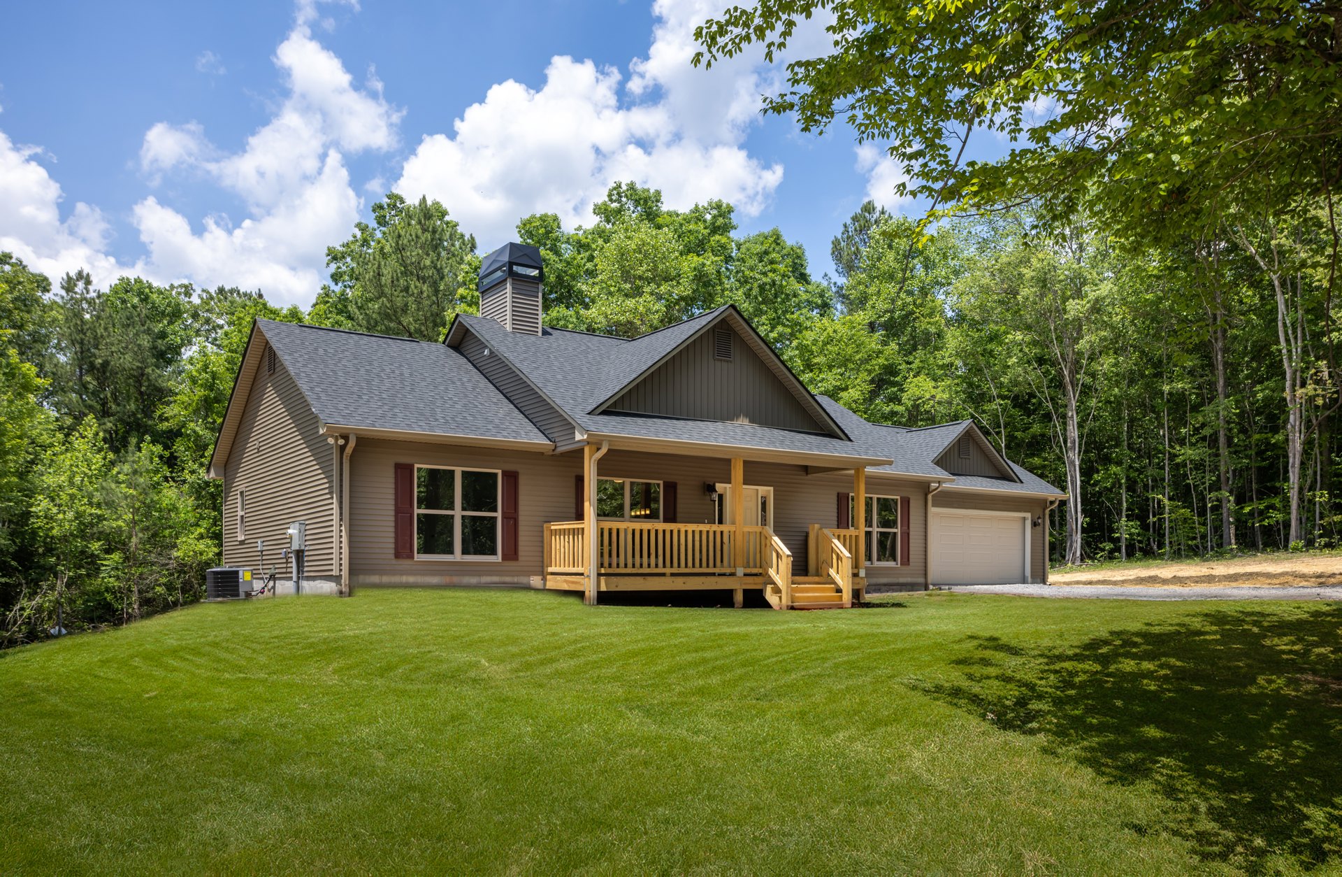 Two-story home with white-framed windows, attached garage, covered front porch featuring a wooden bench, landscaped lawn, and wooden deck surrounded by trees and plants.