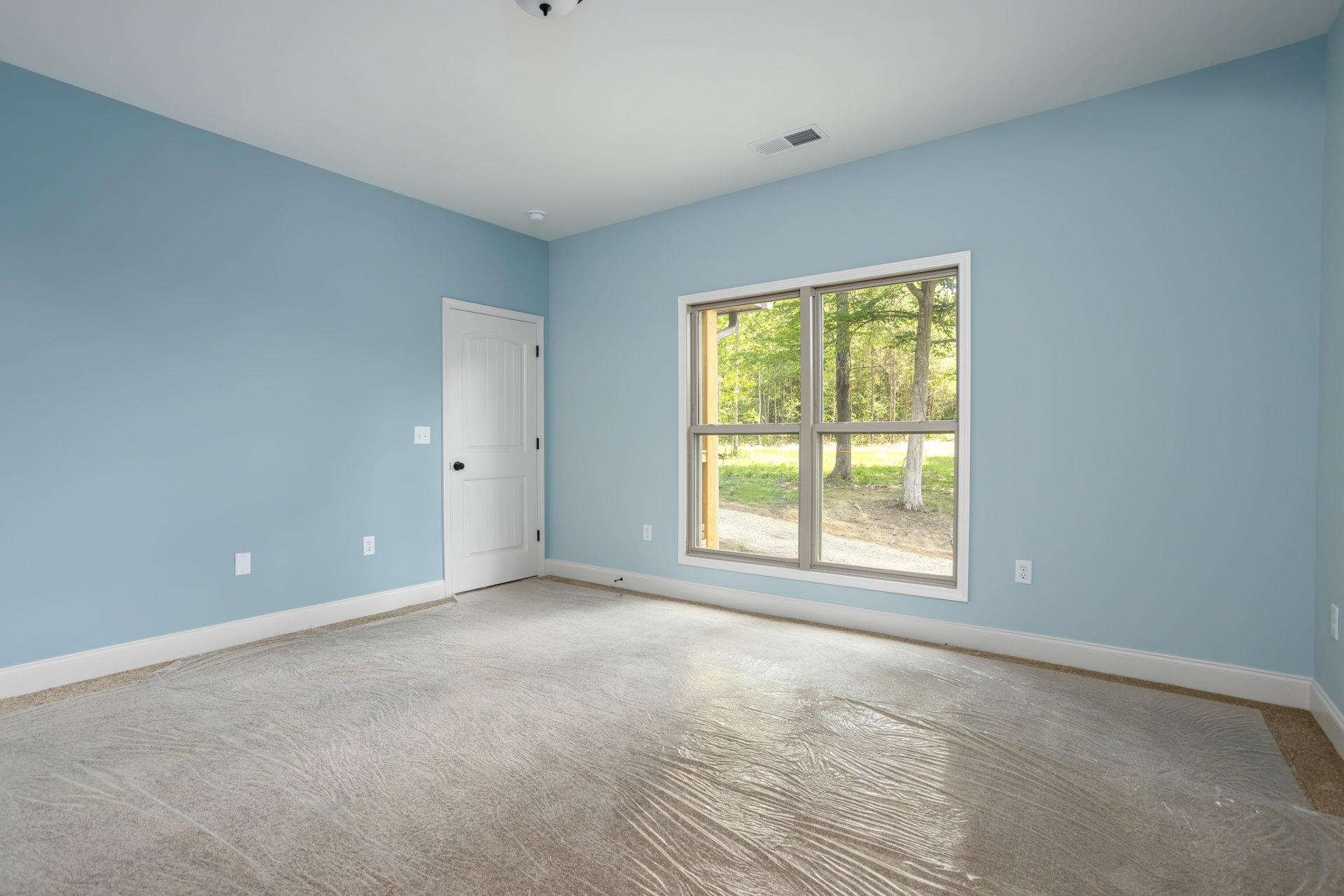 Bedroom with beige carpet, large window overlooking trees, white door with black knob, ceiling vent, and plastic wrap covering part of the floor