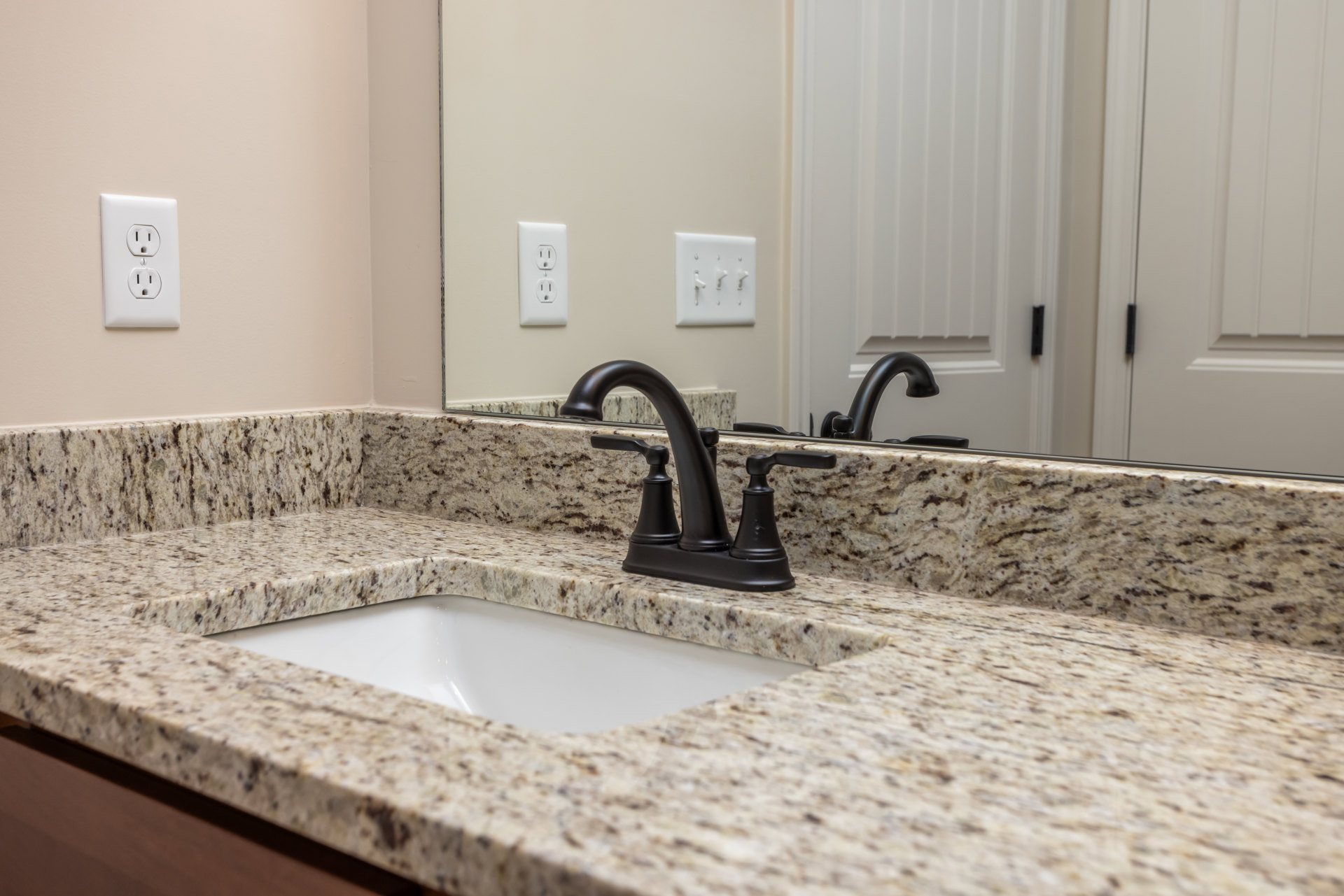 Black faucet mounted on marble countertop with rectangular sink, white tile backsplash, and partial view of wall outlet and door with black handle