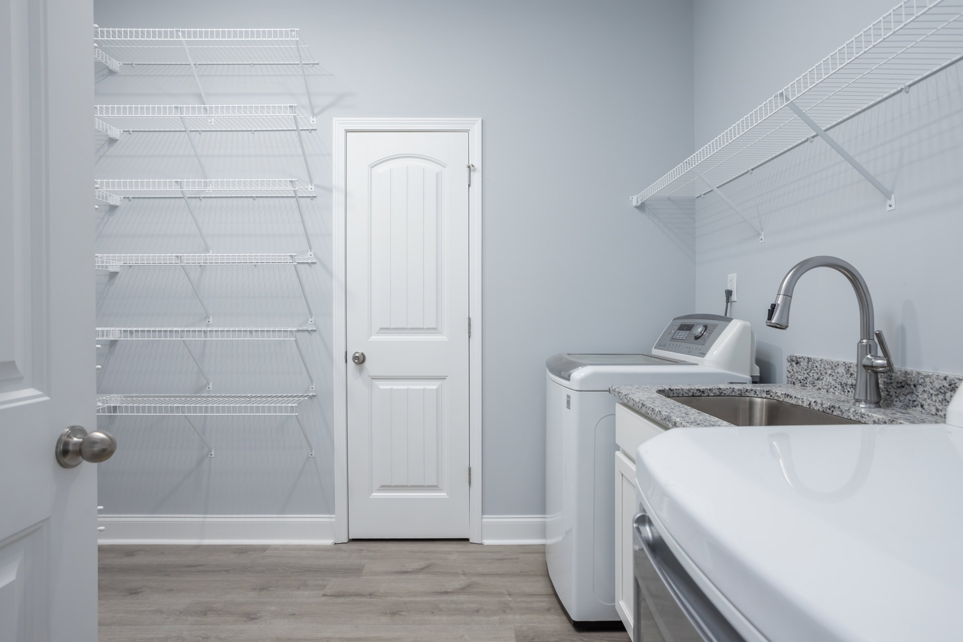 Laundry room featuring white cabinetry, white countertop with stainless steel sink and faucet, white shelving with metal rods, and a white door with a silver knob.