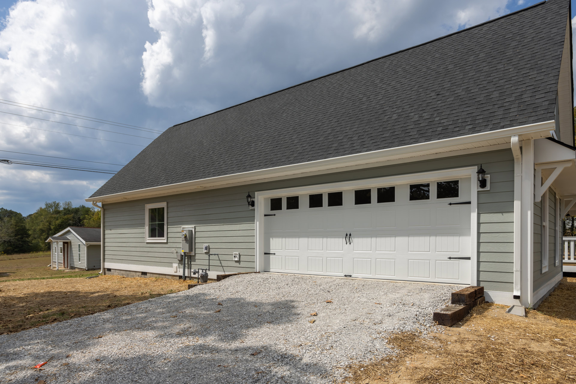 Detached garage with white framed window, gravel driveway, gray roof, and grassy lawn under cloudy sky