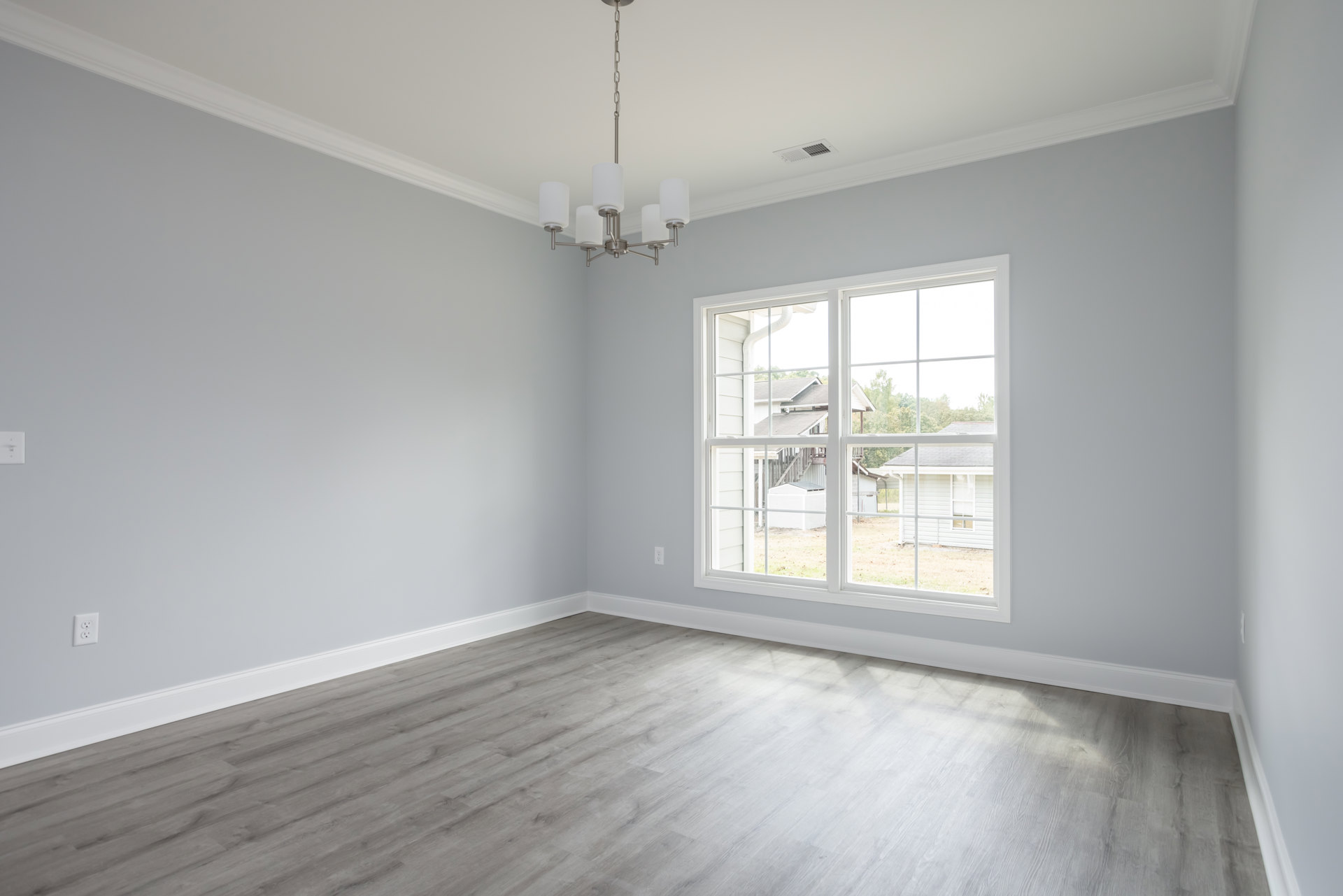 Sunlit room with wide wood plank flooring, large window overlooking yard and neighboring house, white plaster walls, ceiling light fixture, and decorative molding.