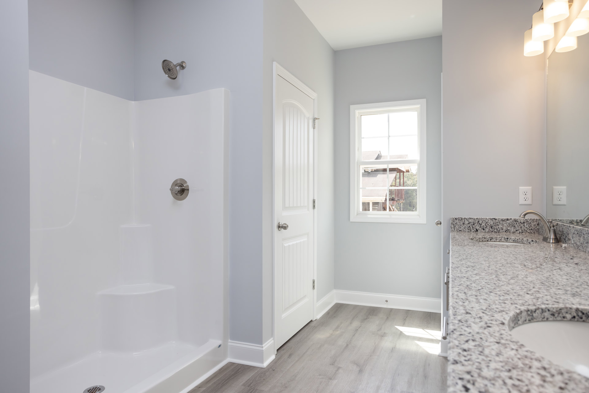 Modern bathroom featuring a tiled walk-in shower with round shower head, double sinks set in a light countertop, white door with silver knob, and window overlooking neighboring
