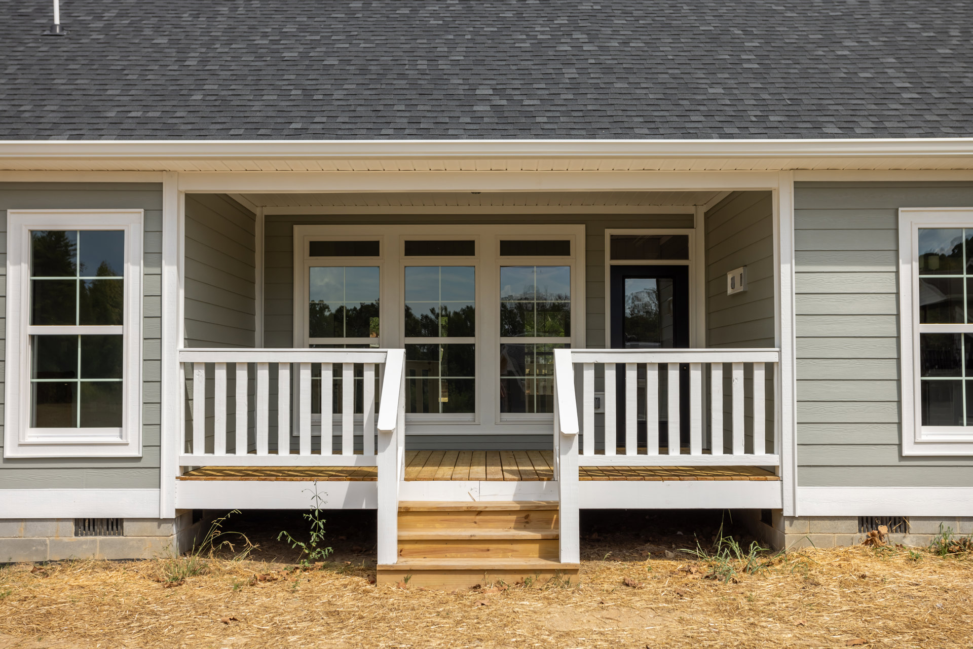 White front porch with wooden deck and railing, white-framed window, wooden steps leading to straw-covered ground, light siding exterior, green grass and plants nearby