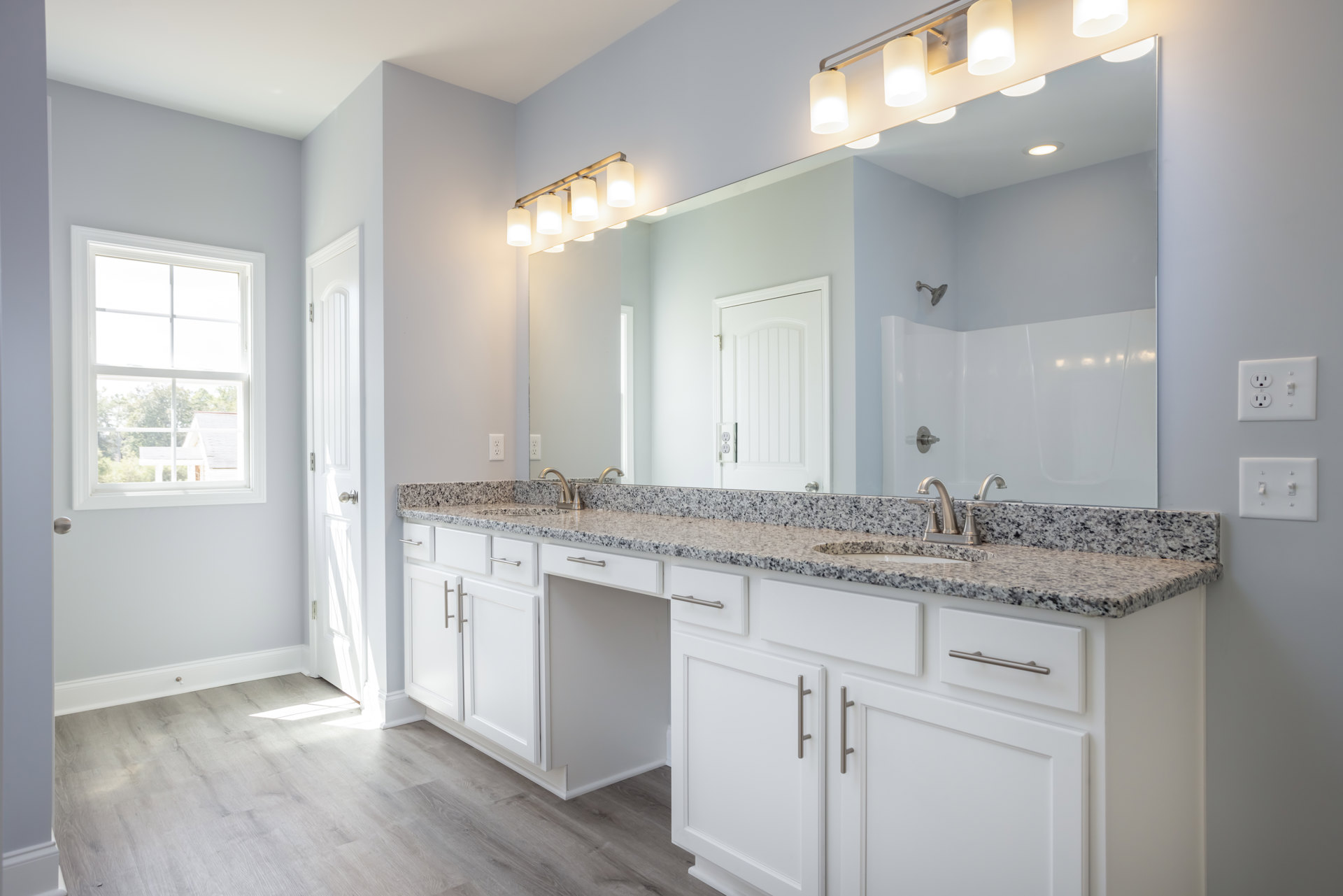 Bathroom with marble countertop, large wall mirror, white cabinetry, chrome faucet, tiled backsplash, window overlooking trees and neighboring house, white door with power outlet