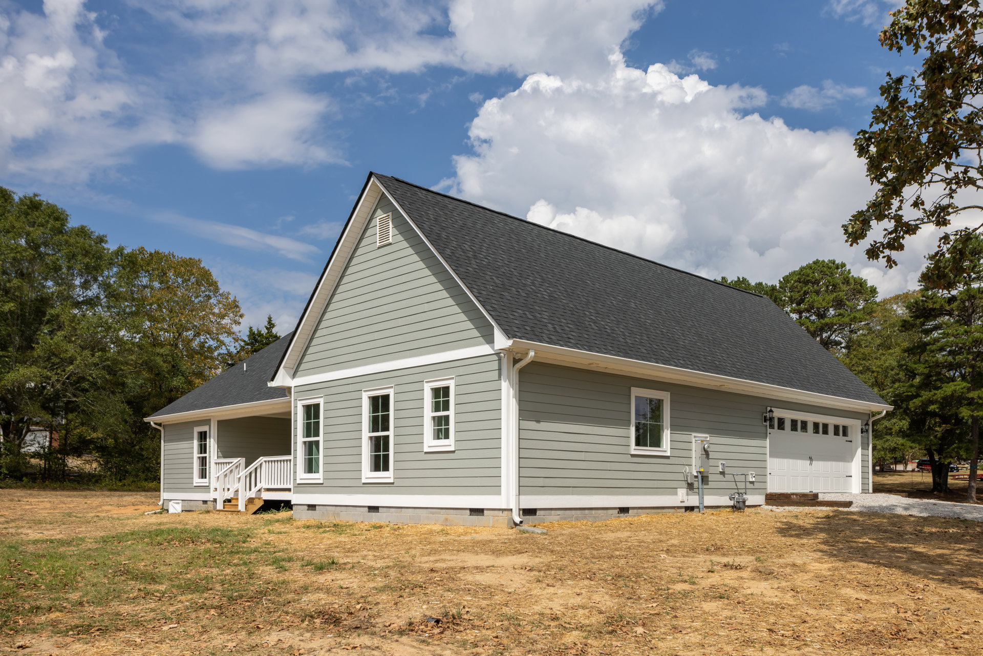 Two-story farmhouse with gray siding, white trim, attached garage, covered front porch with white railing, manicured lawn, and mature trees under partly cloudy sky