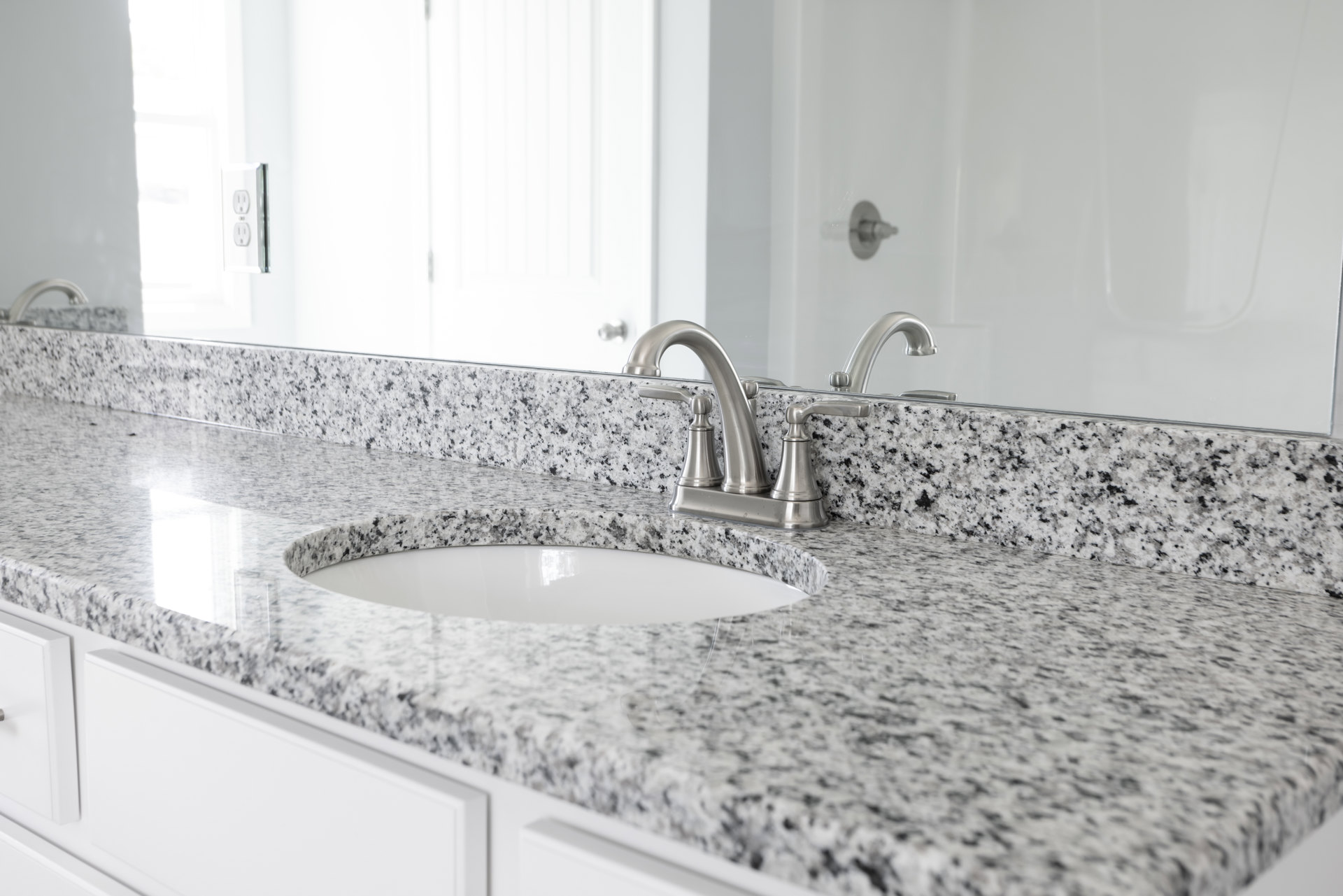 White undermount sink with chrome faucet set in granite countertop, surrounded by light tile backsplash and bathroom accessories