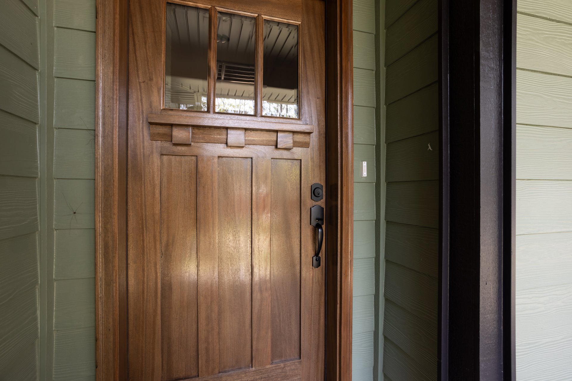 Wooden door with vertical glass panel and black handle, white wood paneling with black trim, window with vent visible in background