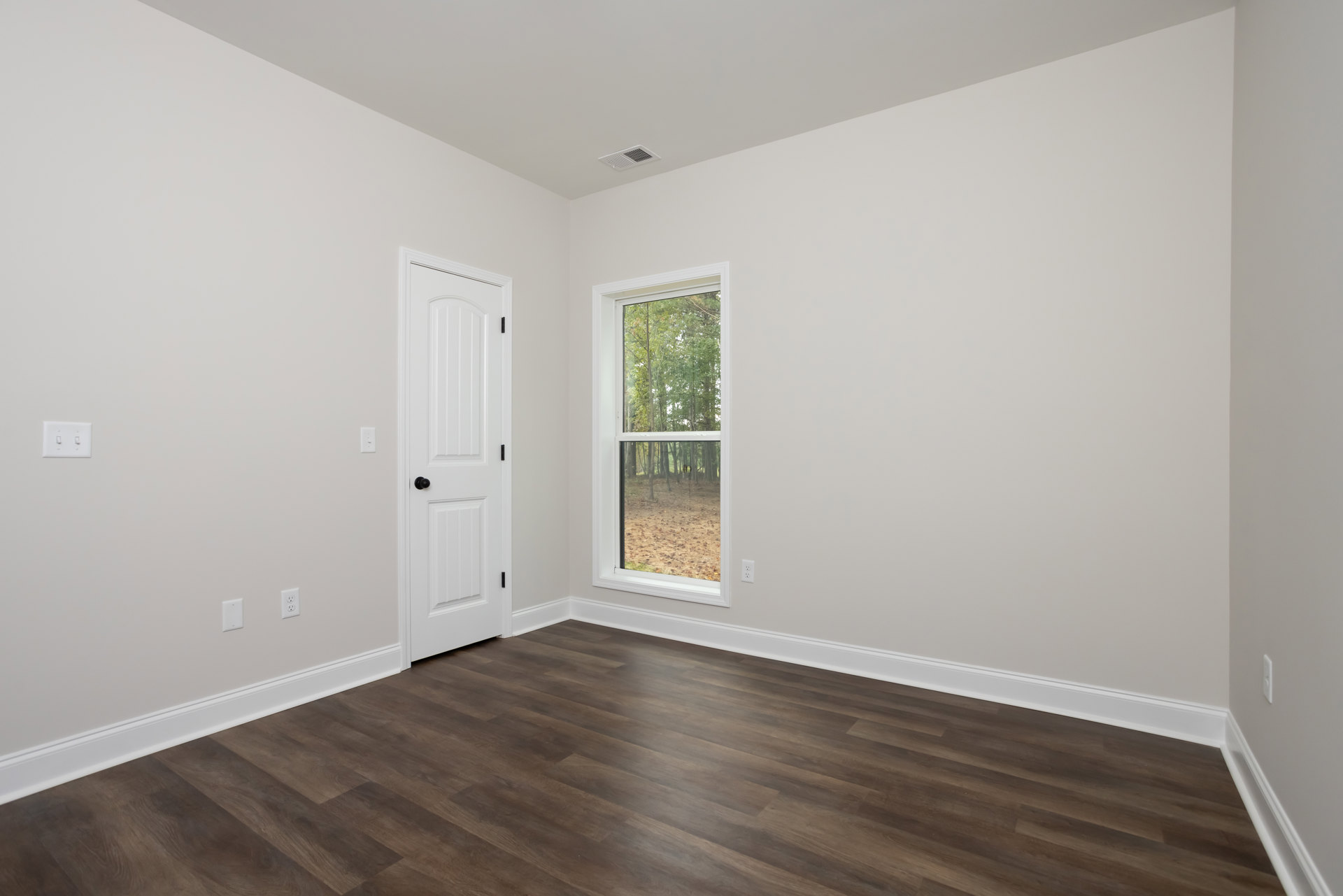 Wood flooring with white trim, white door featuring a black knob, two-switch light plate, large window showing green trees outside
