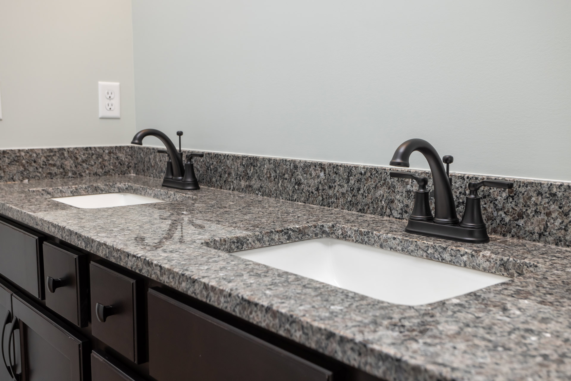 White bathroom sink with matte black faucet set on speckled granite countertop, white tile backsplash, and electrical outlet visible on wall.