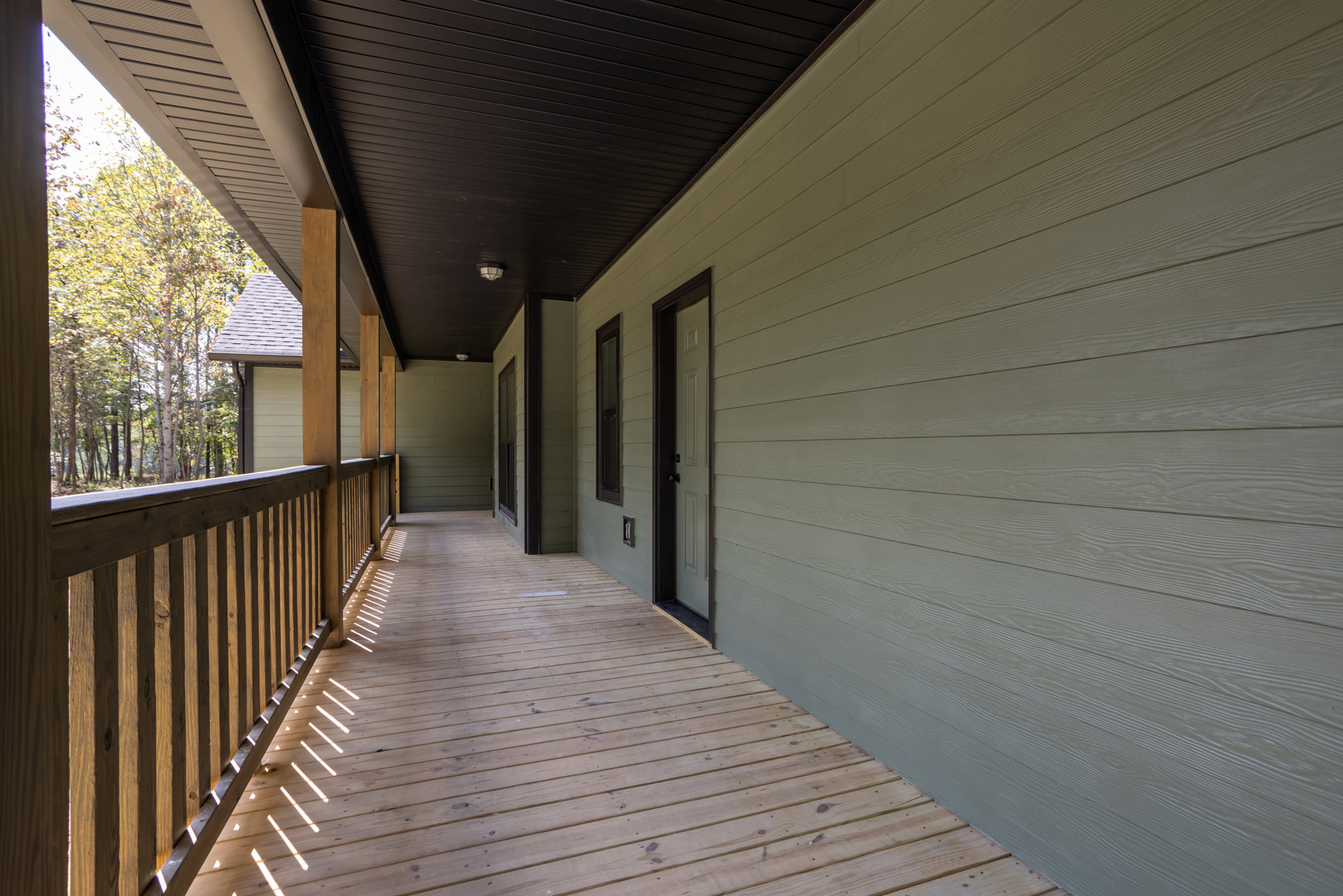 Wood porch with horizontal deck boards, black-handled door, wood railing, and partial roof; sunlight highlights natural finishes, trees visible beyond exterior.