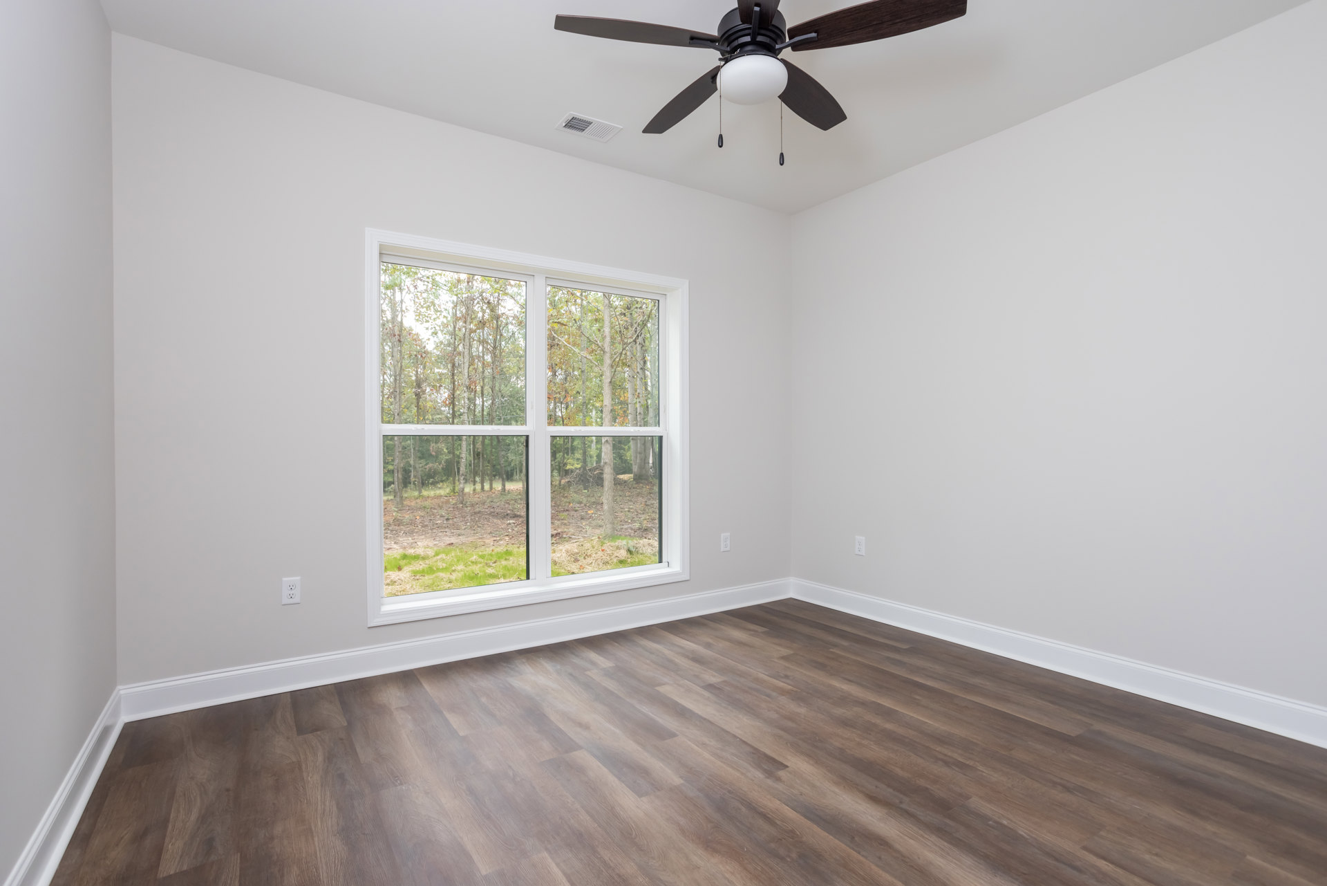 Ceiling fan with light fixture above wood flooring, large window with view of trees, white plaster walls