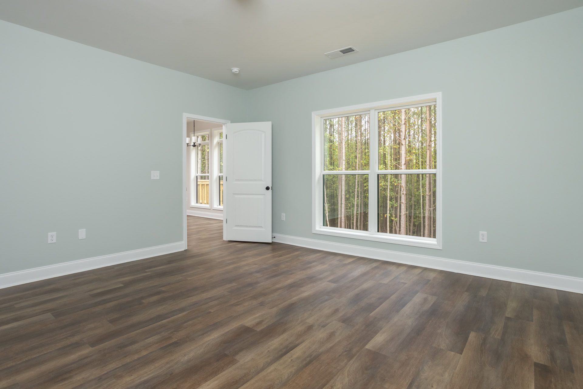 Room with light brown hardwood floor, open white door with black knob, window showing trees outside, white plaster walls