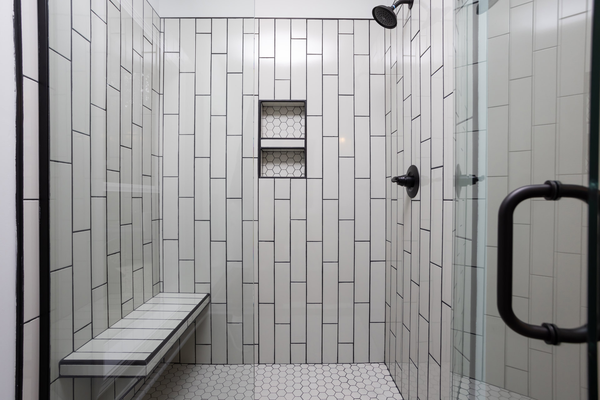 White tile shower with built-in bench, chrome shower head, black and white hexagon shelf, black accent border, and handle on white wall