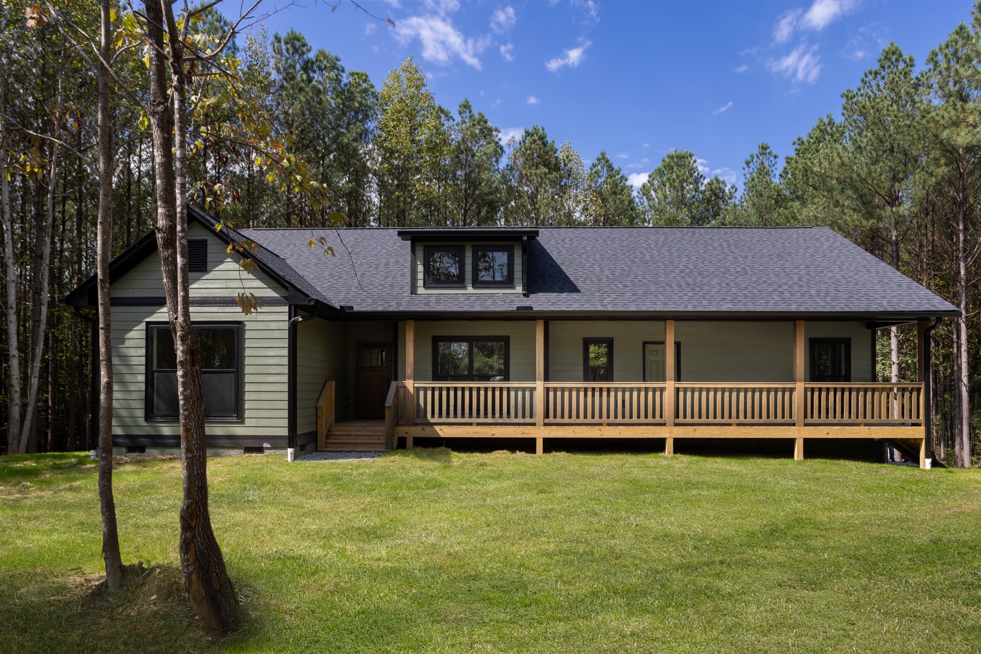 Two-story house with white siding, covered front porch with wooden railing, large windows reflecting leafy tree, manicured grass lawn, cloudy sky overhead