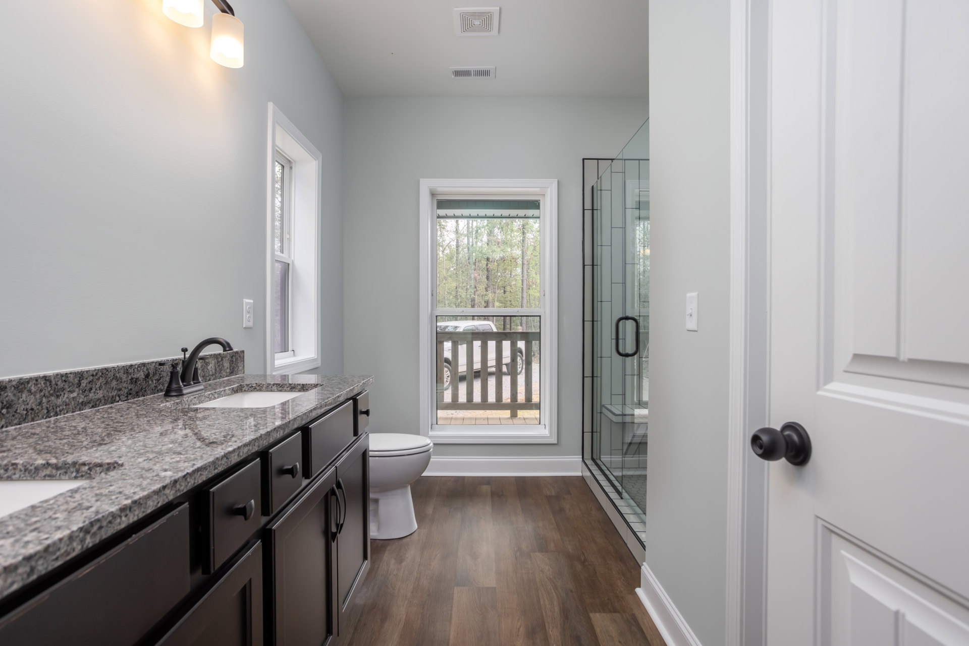 Bathroom featuring a glass-enclosed shower, white toilet with seat down, light-colored tile flooring, and a window with a white frame overlooking a parked car and wooden fence