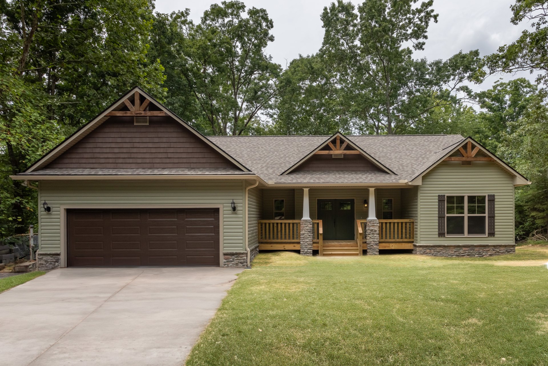 Two-story house with white siding, multi-pane windows, attached garage with paneled door, concrete driveway, wooden porch with railing, grassy lawn, mature trees in background