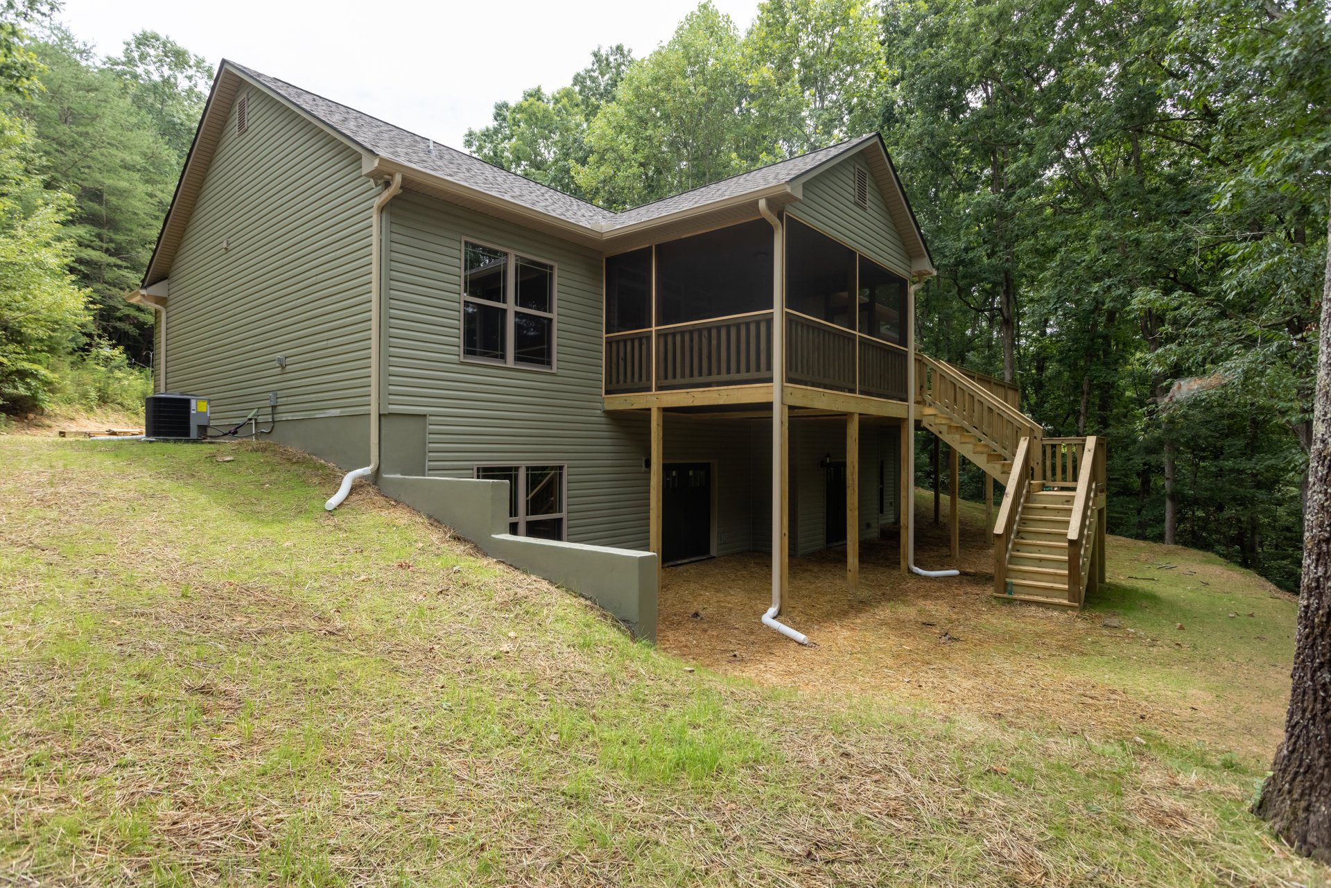 Wooden deck with outdoor stairs leading to a green lawn, grey air conditioner unit beside the house, large window reflecting trees, white pipe on ground, light-colored siding and
