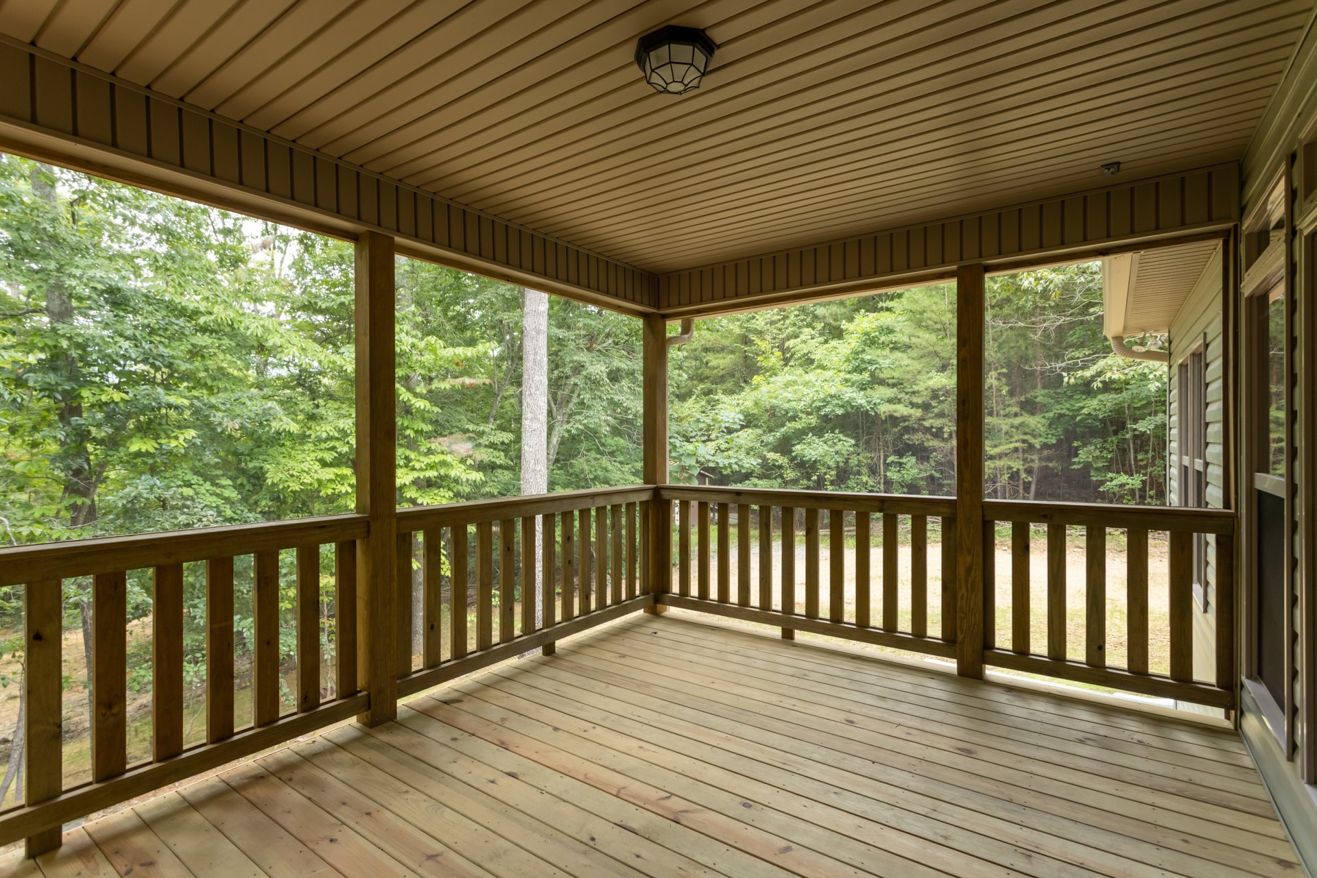 Wooden deck with railing overlooking leafy trees, ceiling light fixture above, shaded outdoor space