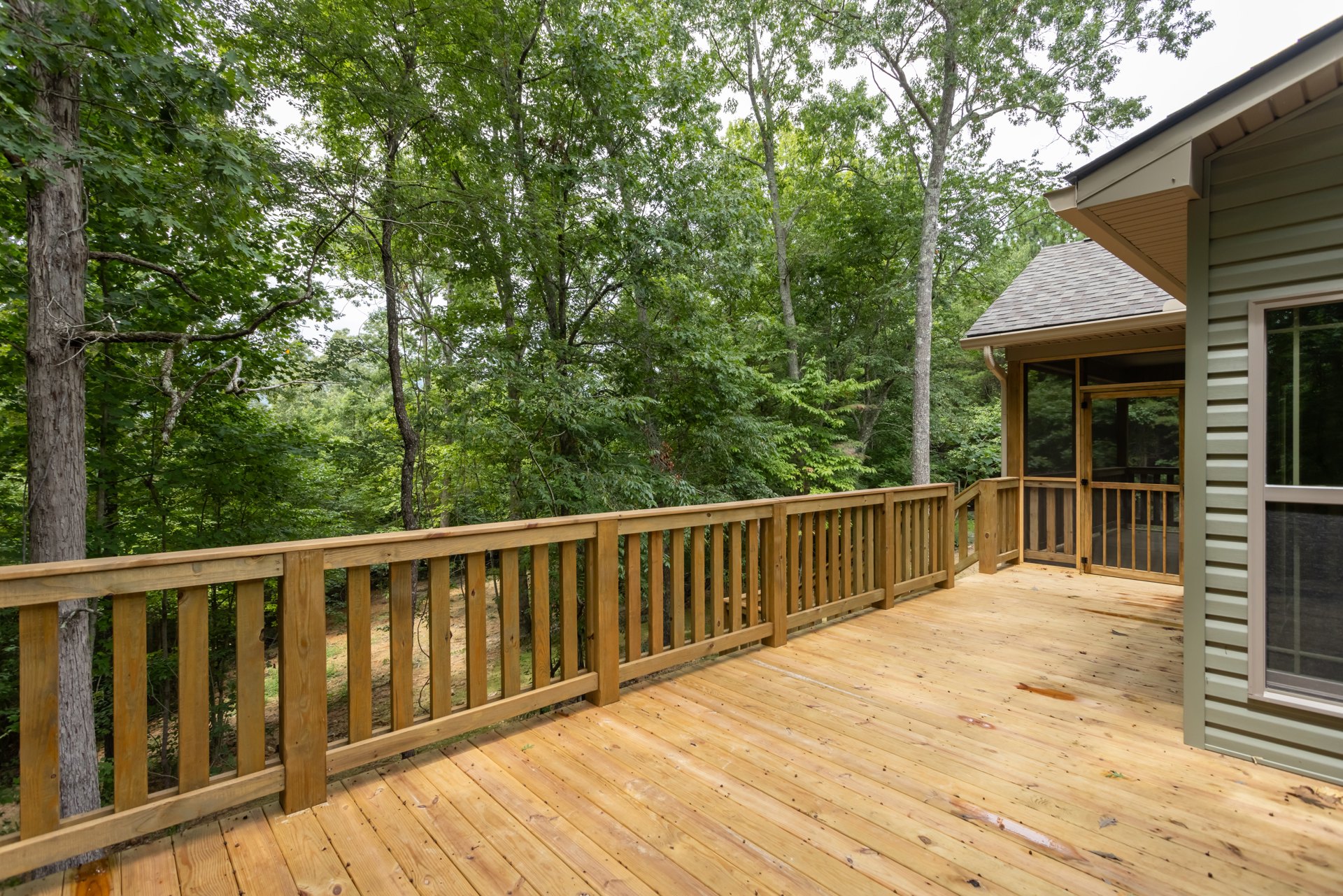 Wooden deck with horizontal wood railing, screen door, and leafy trees in the background