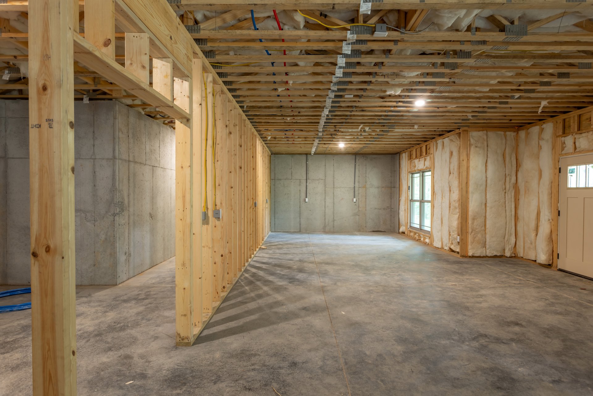 Concrete floor and wall, exposed wood ceiling beams, white-framed window with paper taped to glass, unfinished wood framing with visible wiring