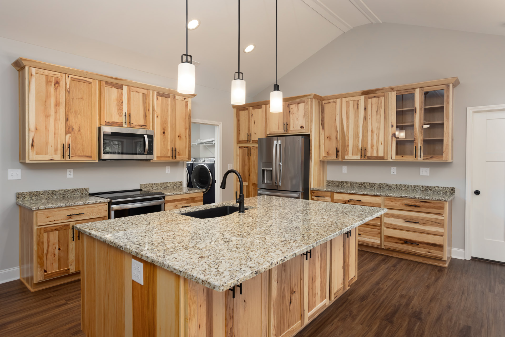 Spacious kitchen featuring a large central island with built-in sink, double-door stainless steel refrigerator, wood cabinetry surrounding a microwave near a window, white