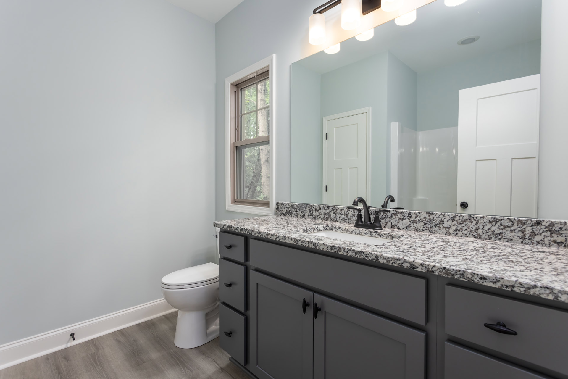 Bathroom with white toilet, rectangular sink set in white cabinetry, square panel door with black hinge, wall-mounted mirror, tile flooring, window showing green trees outside