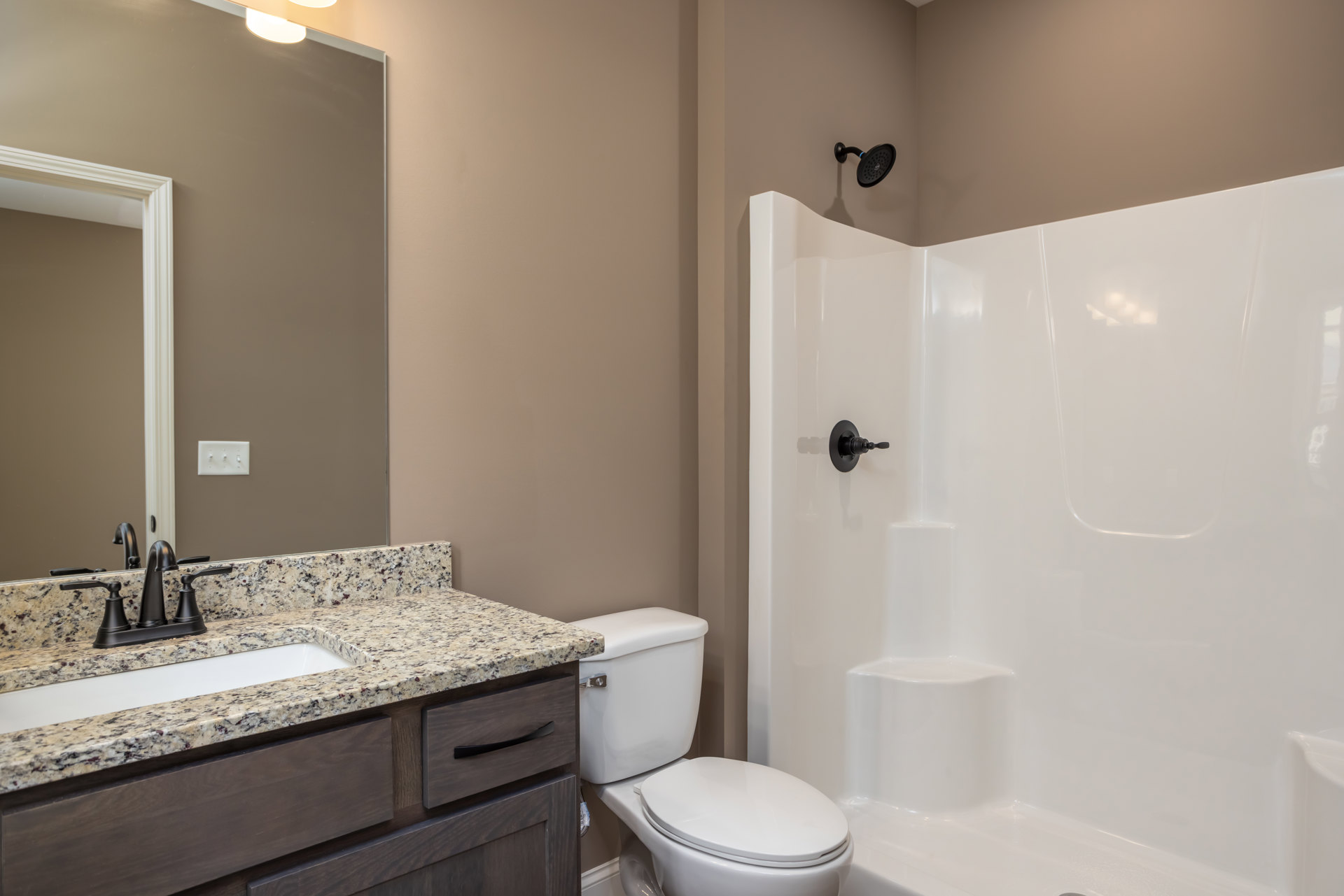 Bathroom with white toilet, marble countertop vanity with sink and drawers, white shower featuring black handle, light tile flooring, and neutral wall finishes.