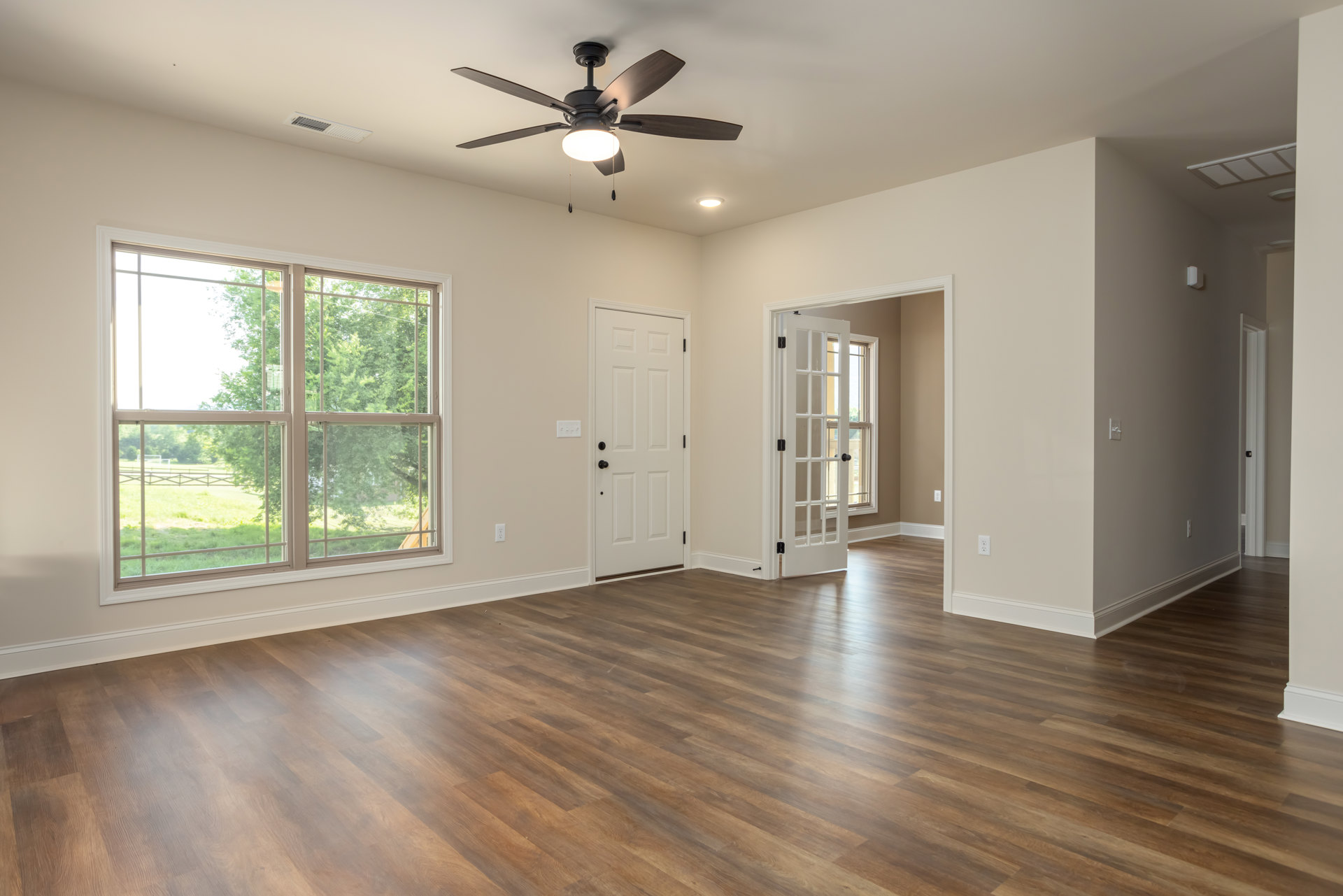 Ceiling fan with light fixture, hardwood floor, white walls, window showing trees outside, white door with black knobs, glass-paneled door with black handle