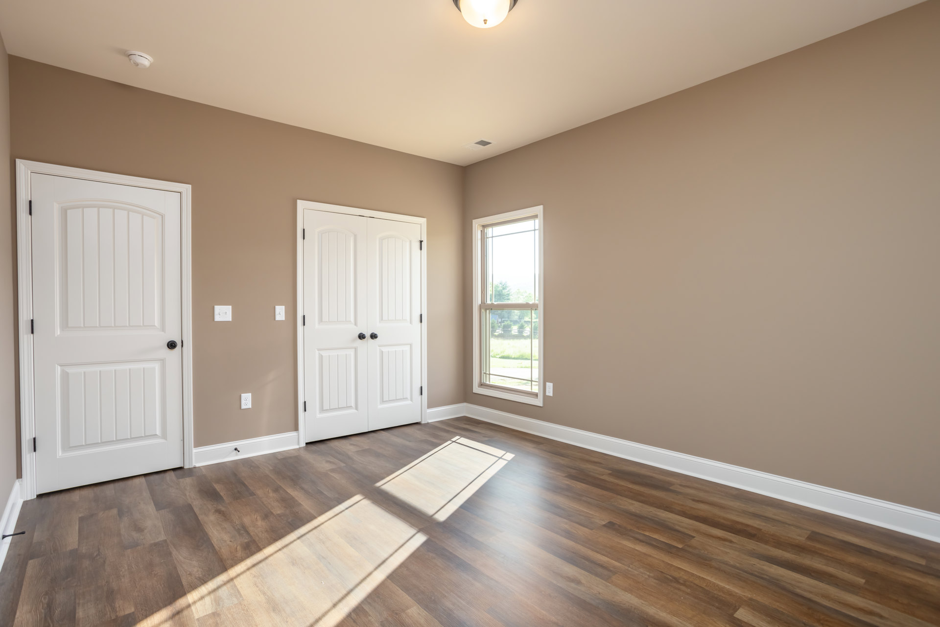 Hardwood floor room with white double doors featuring black knobs, large window overlooking trees and fence, white rectangular object placed on floor